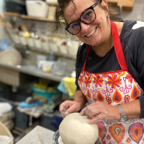 A smiling woman wearing glasses and a colorful floral apron works on a pottery wheel, shaping a large round clay piece in a workshop.