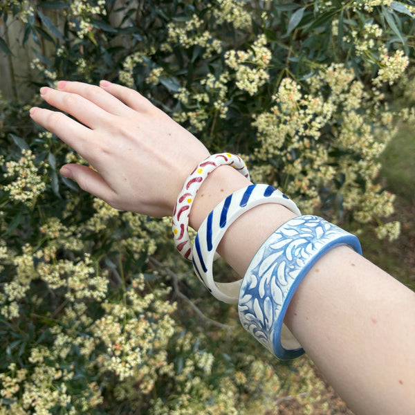 A person's hand and forearm wearing three patterned porcelain ceramic bangles, posed in front of a leafy bush with small pale flowers.