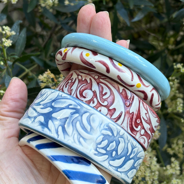 A hand holding a stack of five colourful porcelain ceramic bangles outdoors, with green foliage in the background. The bangles feature various patterns including light blue, red and white scrollwork, blue and white floral motifs and blue and white stripes.