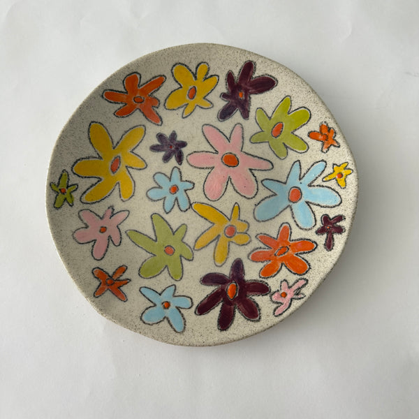 A round ceramic plate with a speckled beige glaze, decorated with a colorful pattern of hand-drawn flowers in yellow, orange, pink, blue, green, and purple, photographed from directly above on a white background.