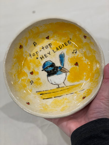 A handmade ceramic bowl held in a hand, featuring a yellow and cream glaze with a painted blue and white bird, small red hearts, and the text 'tap-tap HEY LADIES'.