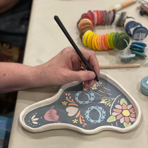 A hand holding a fine paintbrush applies paint to a decorative ceramic tray with a floral pattern on a light-colored table. Spools of thread and small jars of paint are visible in the background.
