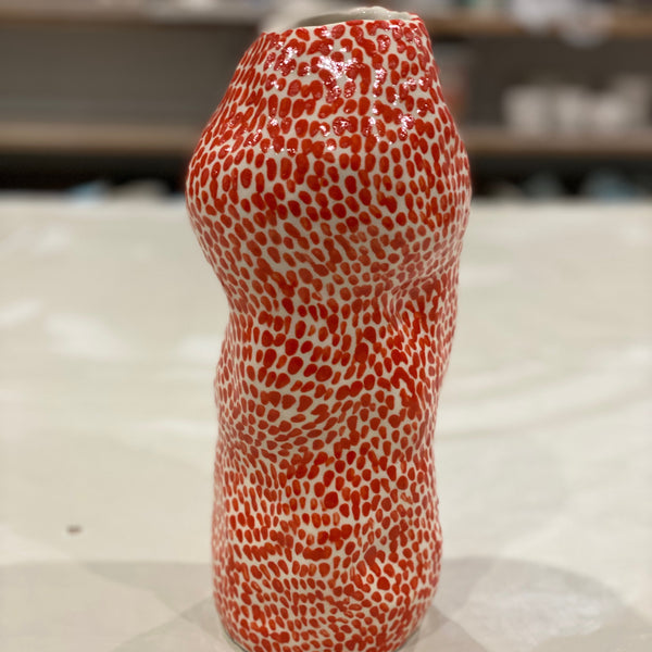 A tall, organic-shaped ceramic vase with a glossy red and white spotted pattern, standing on a light-colored surface with a blurred workshop background.