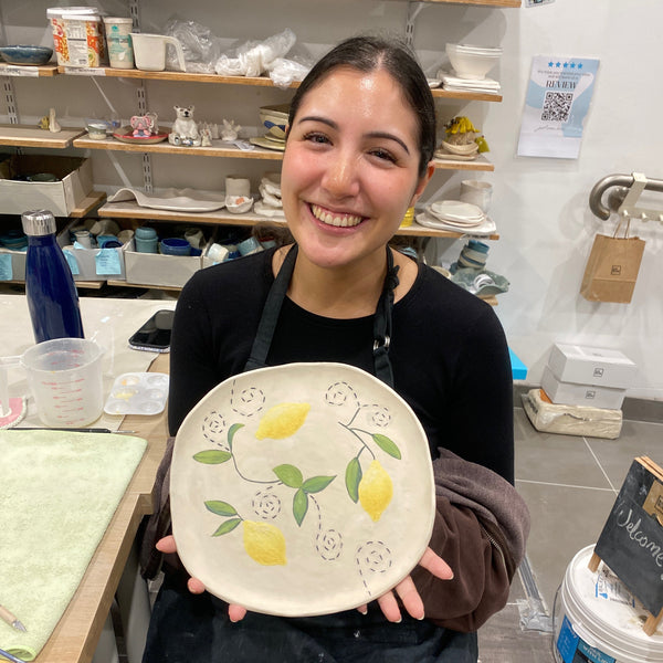 A smiling woman in a black apron holds up a hand-painted ceramic plate decorated with yellow lemons and green leaves, set against a pottery studio background with shelves of pottery.