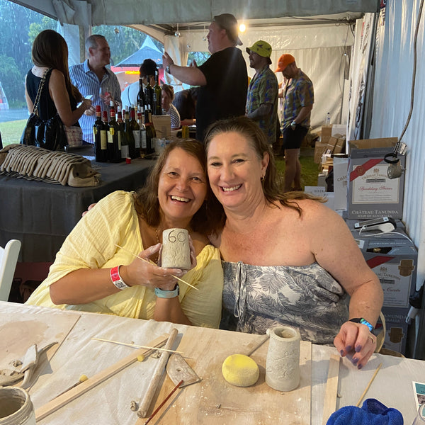 Two smiling women posing together at a pottery workshop at a Wine Event making  ceramics  with clay tools, clay, outdoors, under a tent.