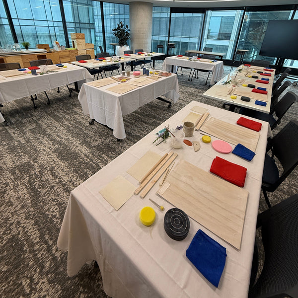 A long table in a modern indoor space is covered with craft or art supplies, including colorful rectangular pieces, wooden sticks, small cups, and a black circular object, photographed from an angled overhead perspective.