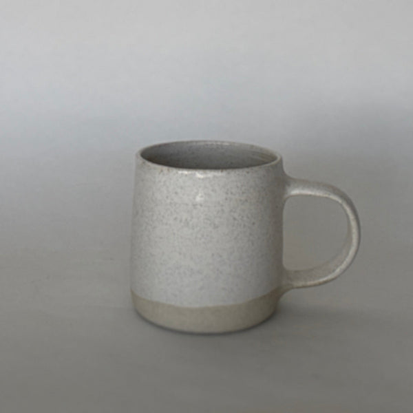A speckled ceramic mug with a handle, sitting on a neutral gray surface against a matching gray background.