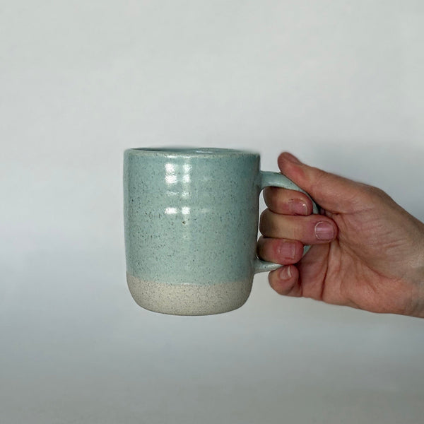 A hand holding a ceramic mug with a two-tone glaze, featuring a light teal upper half and a natural off-white lower half, set against a plain light gray background.