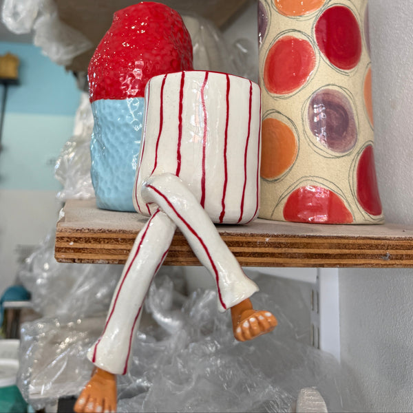 A ceramic art piece depicting a character sitting on a wooden shelf, wearing red-and-white striped pajamas with bare feet, next to a tall cylindrical vase with colorful polka dots.