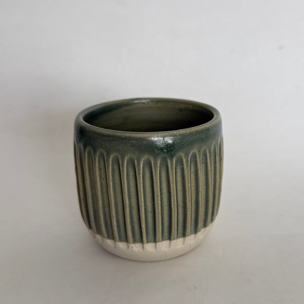 A small handmade ceramic cup with a ribbed texture, featuring a dark green glazed upper half and a white unglazed base, set against a plain light gray background.