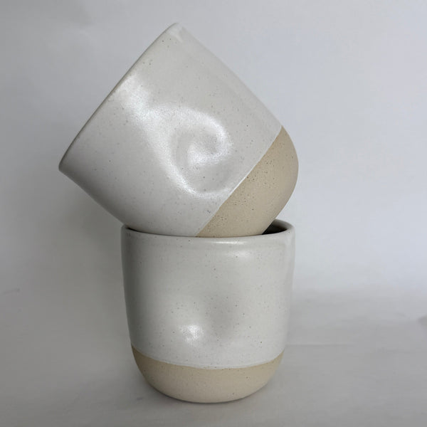 Two handmade ceramic cups stacked on top of each other, featuring a glossy white upper half with a subtle thumb dimple and a matte beige lower half, set against a neutral light gray background.