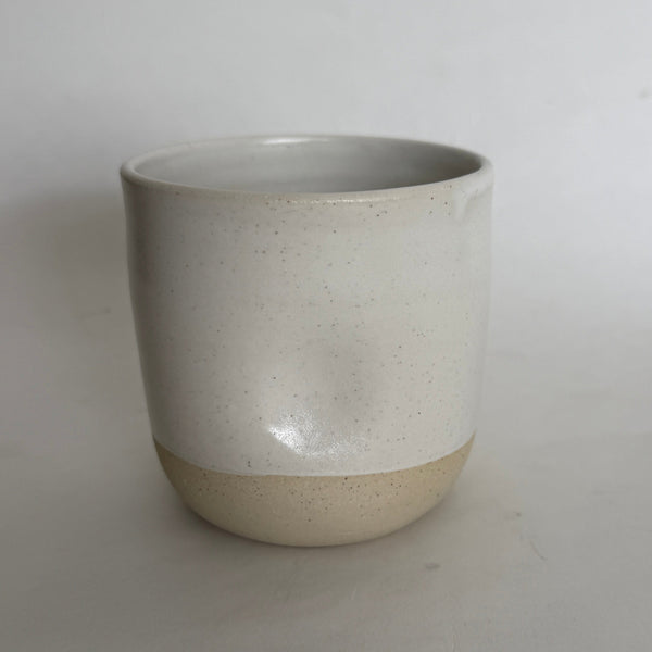 A handmade ceramic cup with a speckled off-white glaze on the upper half and a natural, unglazed beige clay base, set against a plain light gray background.