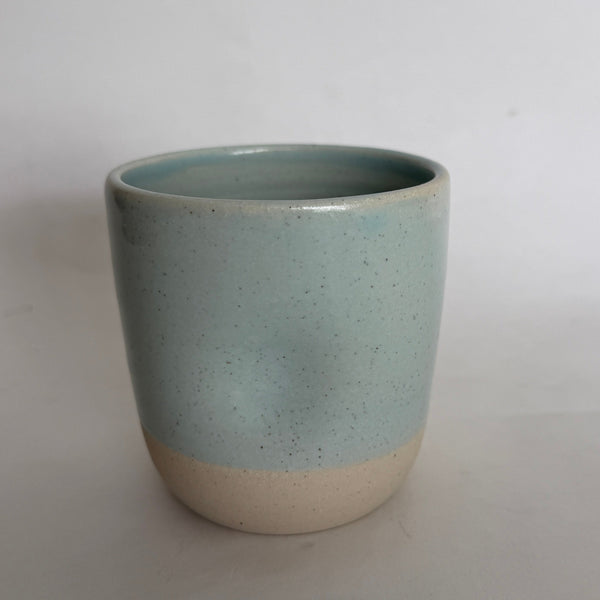 A handmade ceramic cup with a two-tone finish, featuring a speckled light blue-green glazed upper half and a natural beige unglazed lower half, set against a plain light gray background.