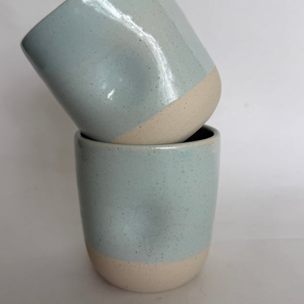 Two handmade ceramic cups stacked together, featuring a speckled light blue glaze on the upper half and a natural beige unglazed finish on the bottom half, set against a plain light gray background.