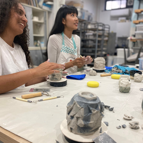 Two women working with clay to create Cups with the Kurinuki method of carving, shaping clay vessels with hand tools and banding wheels in a studio setting.