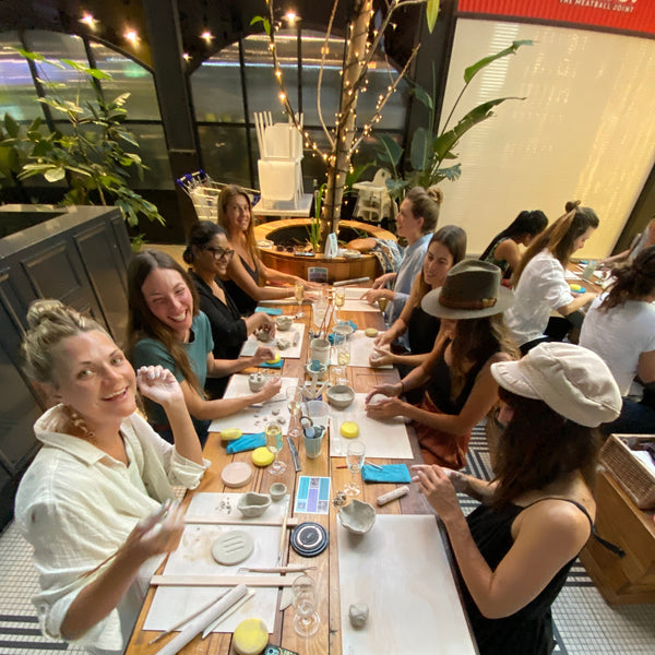 A group of people gathered around a long wooden table in a cafe or workshop setting, engaged in a clay activity with tools. The scene is viewed from above at an angle, showing a busy, collaborative atmosphere.
